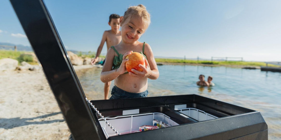 A girl holding an apple and looking at BougeRV's eco-friendly ASPEN PRO fridge near a lake]